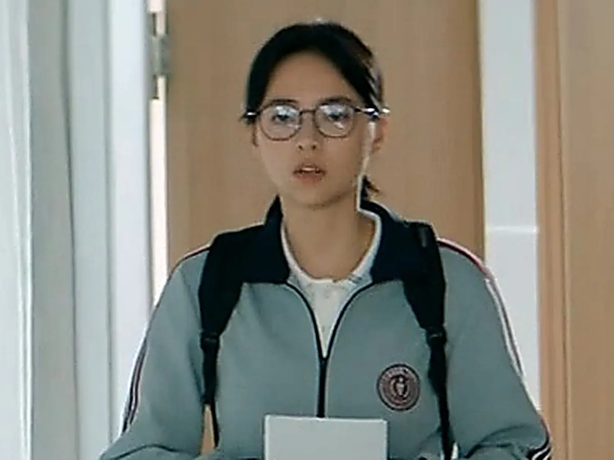 A student wearing round eyeglasses and a grey zip-up jacket with backpack straps holding a notebook