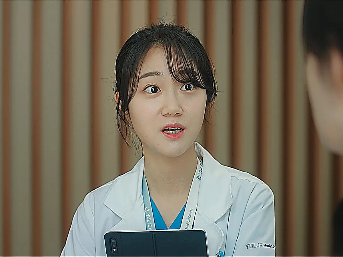 A woman with bangs in a white coat over blue scrubs holding a black tablet