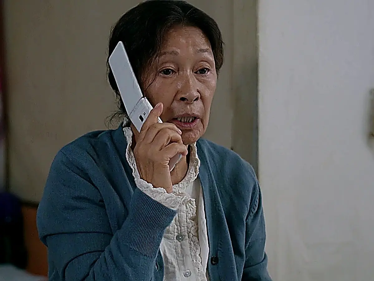 An elderly woman holding a white flip phone to her ear while wearing a blue cardigan over a lace-collared shirt