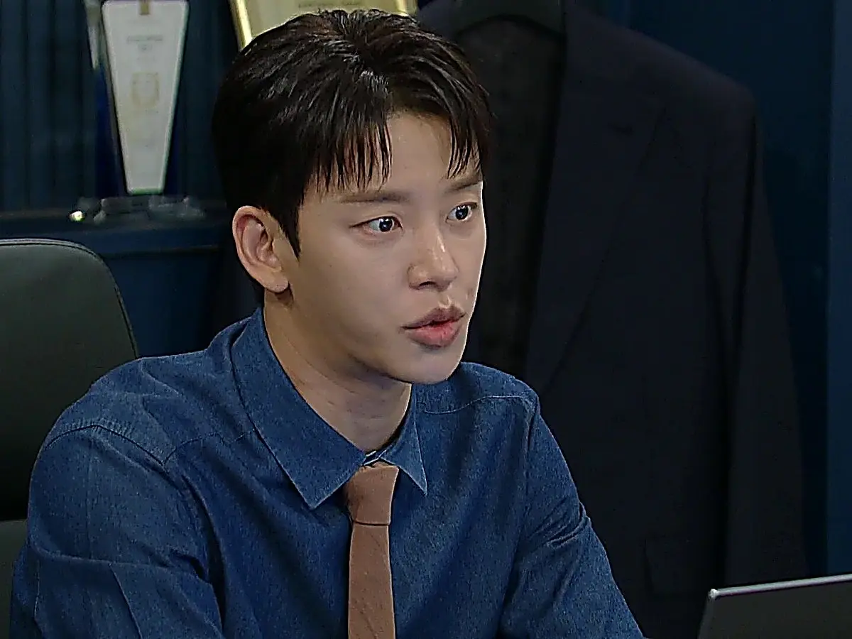 Cha Seok-jin wearing a blue collared button-down shirt paired with a tan knit necktie in his office