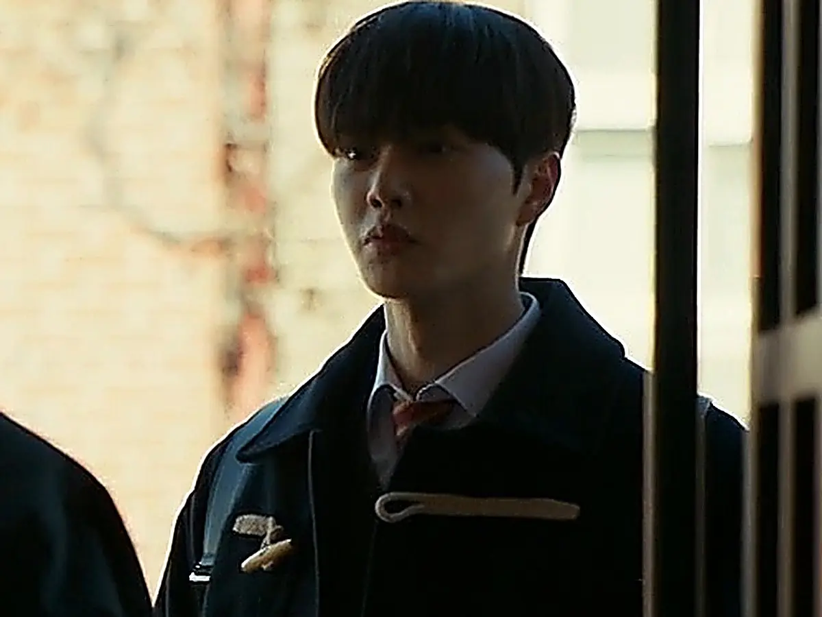 Kang Bi-o standing in a dormitory hallway wearing a navy duffle coat with rope toggles