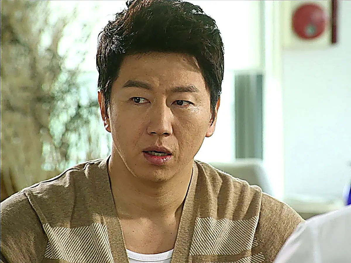 A man in a brown knit shirt over a white undershirt looking forward with a tense, conflicted expression