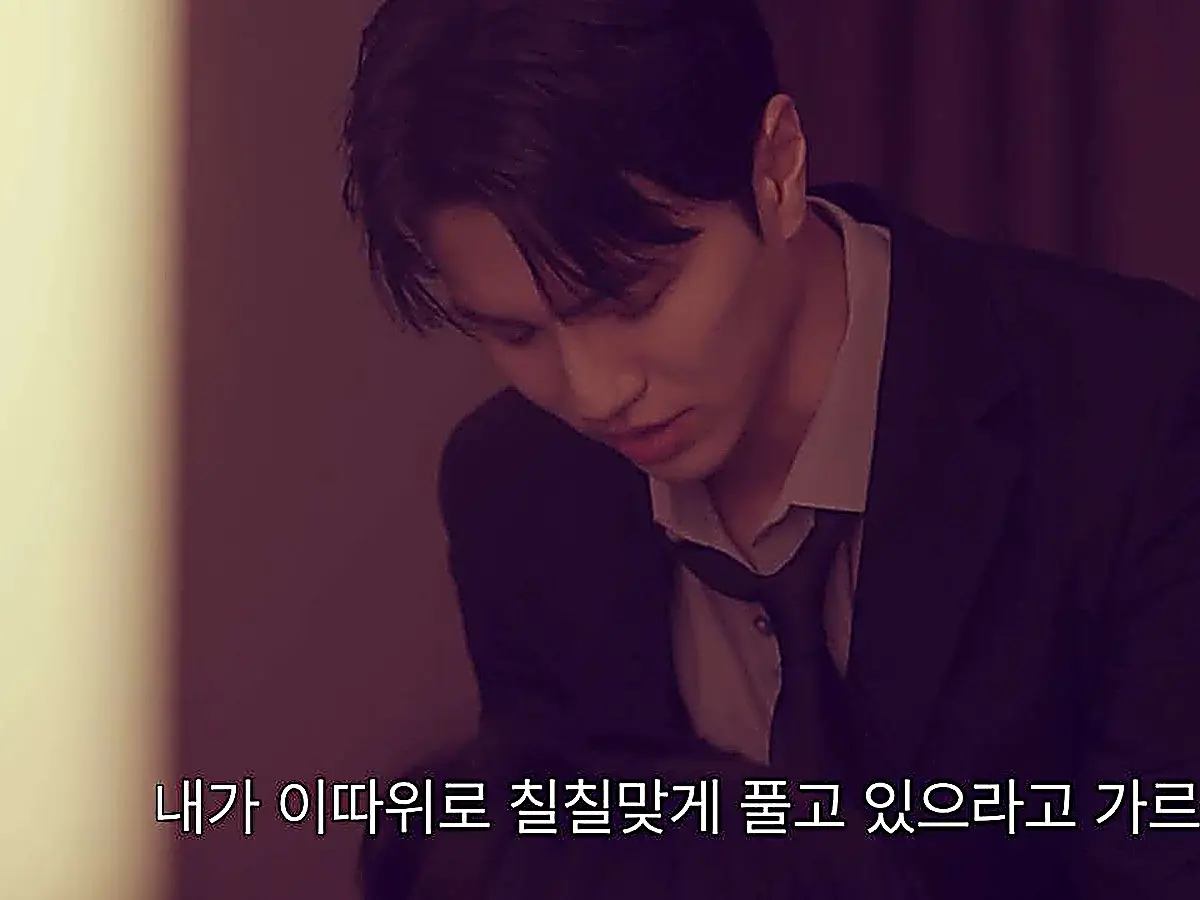 Kang Do-jin looking downward wearing a dark tailored blazer, light dress shirt, and loosened necktie