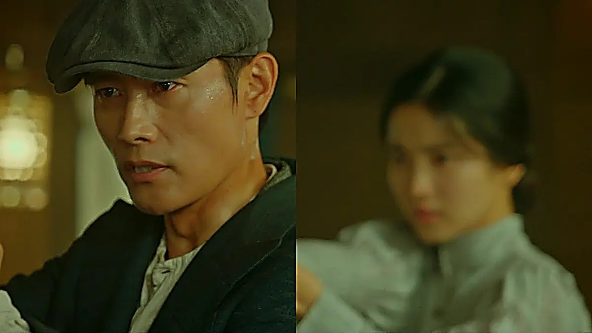 A man in a dark flat cap and overcoat standing in a train compartment with a woman blurred in the background