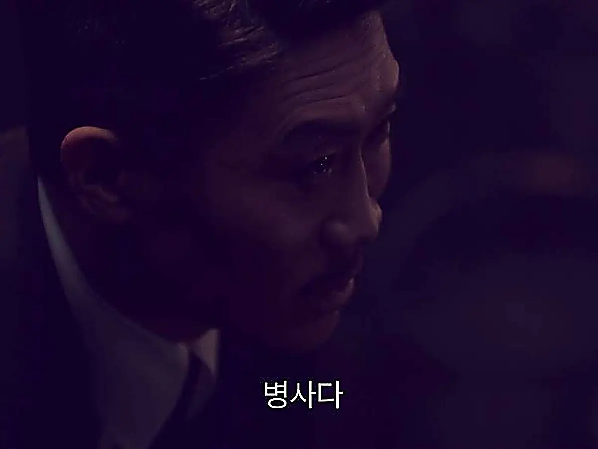 A man with slicked-back hair wearing a dark suit looking downward in profile