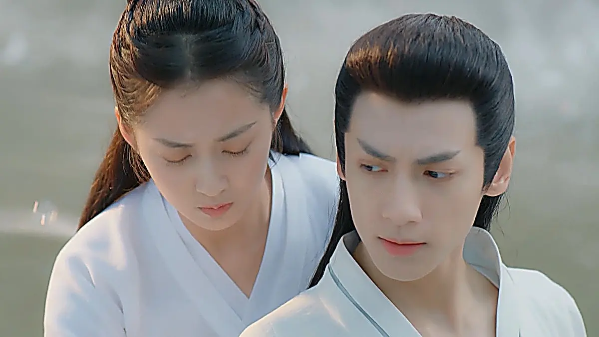 A man and a woman in white robes sitting close together outdoors with misty water in the background