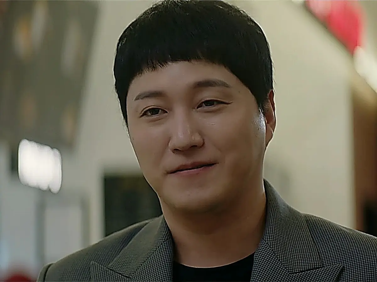 A man with short bangs wearing a grey checkered blazer smiling warmly.