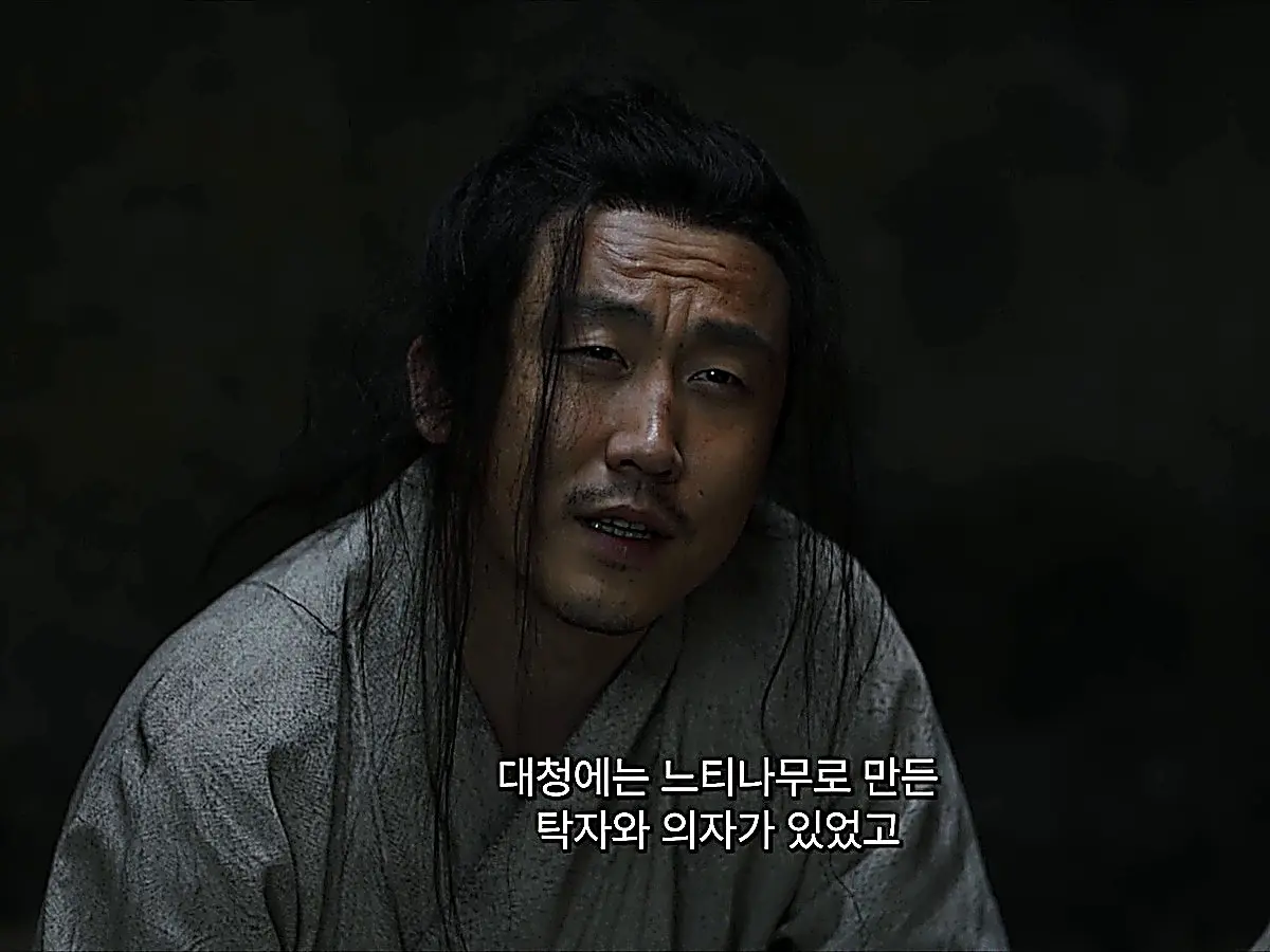 A man with disheveled hair in coarse grey robes speaking inside a dark room