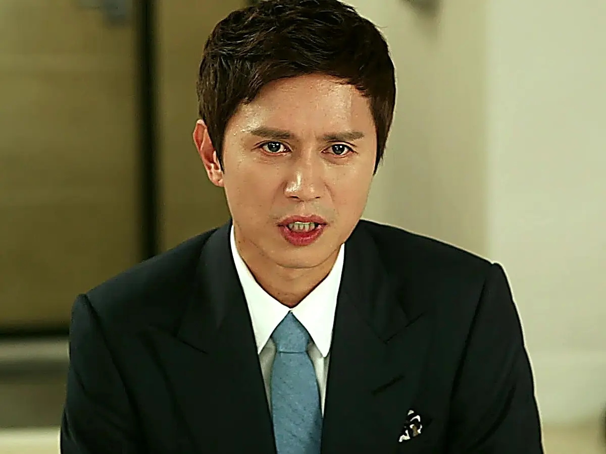 A man in a dark suit jacket and light blue tie looking forward with an earnest expression
