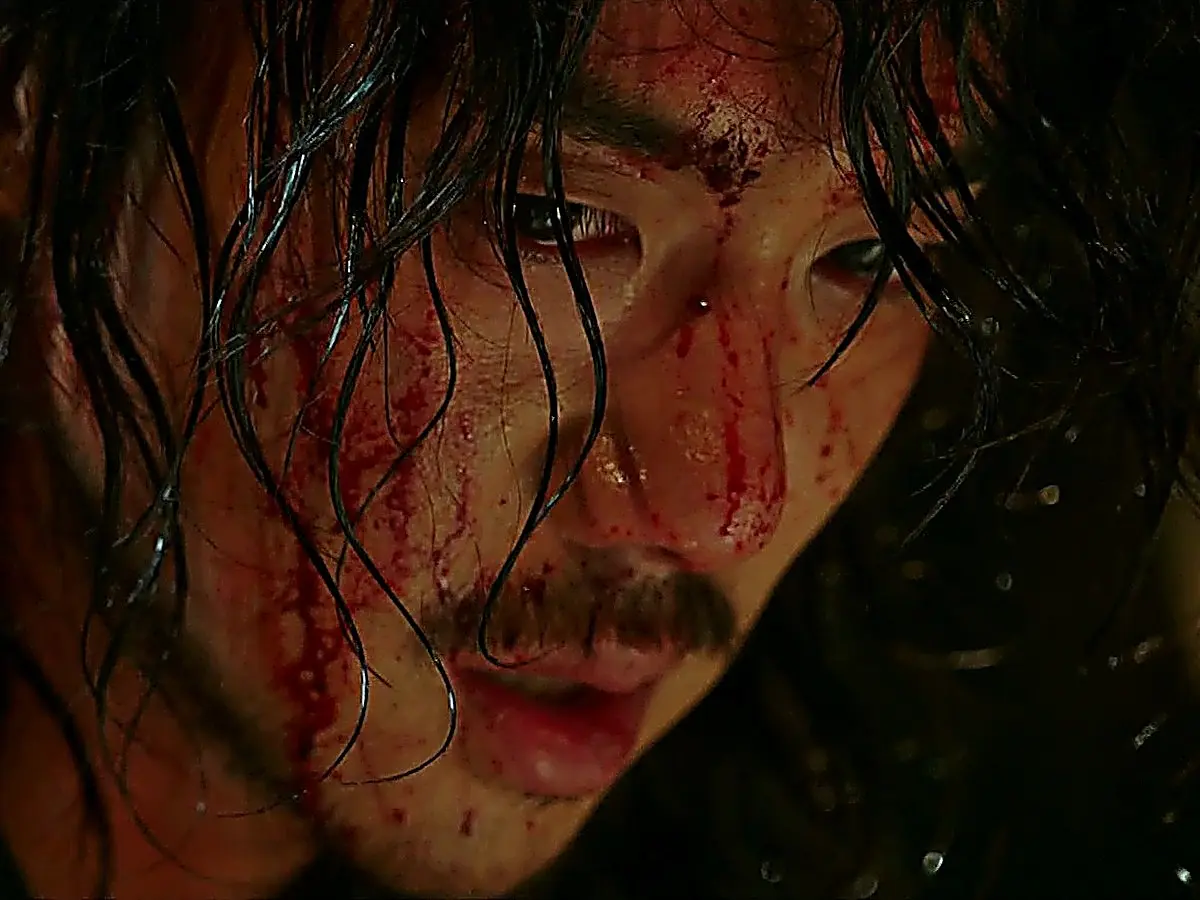 A man with disheveled dark hair and blood on his forehead looking down