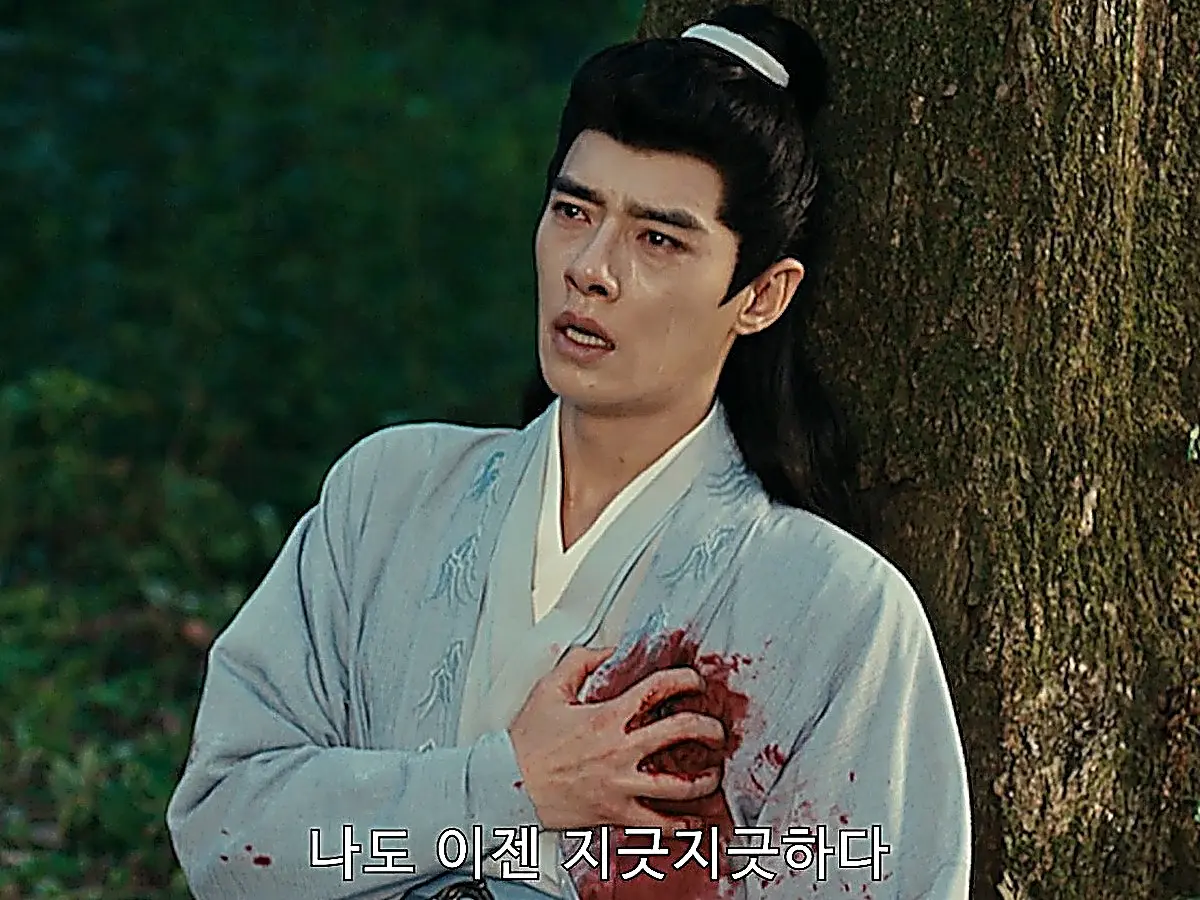 A man in a light blue embroidered robe leaning against a tree trunk with red stains on his chest