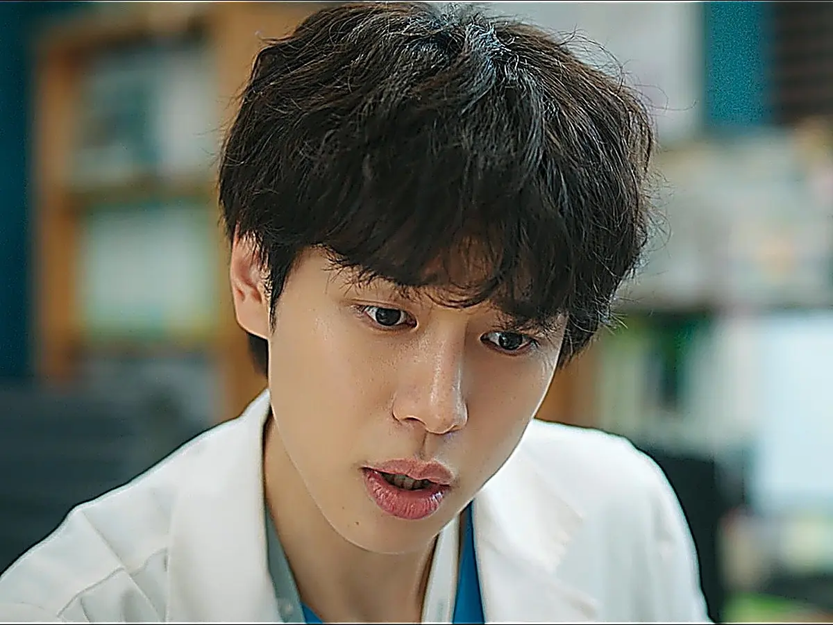 A young man in a white doctor coat and scrubs looking down with an astonished expression