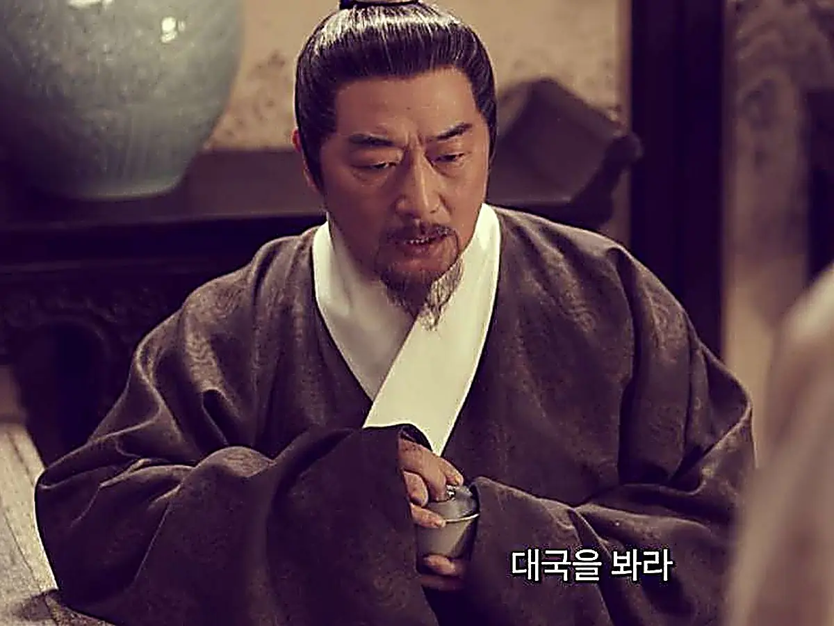 A bearded official in a dark patterned robe holding a small cup