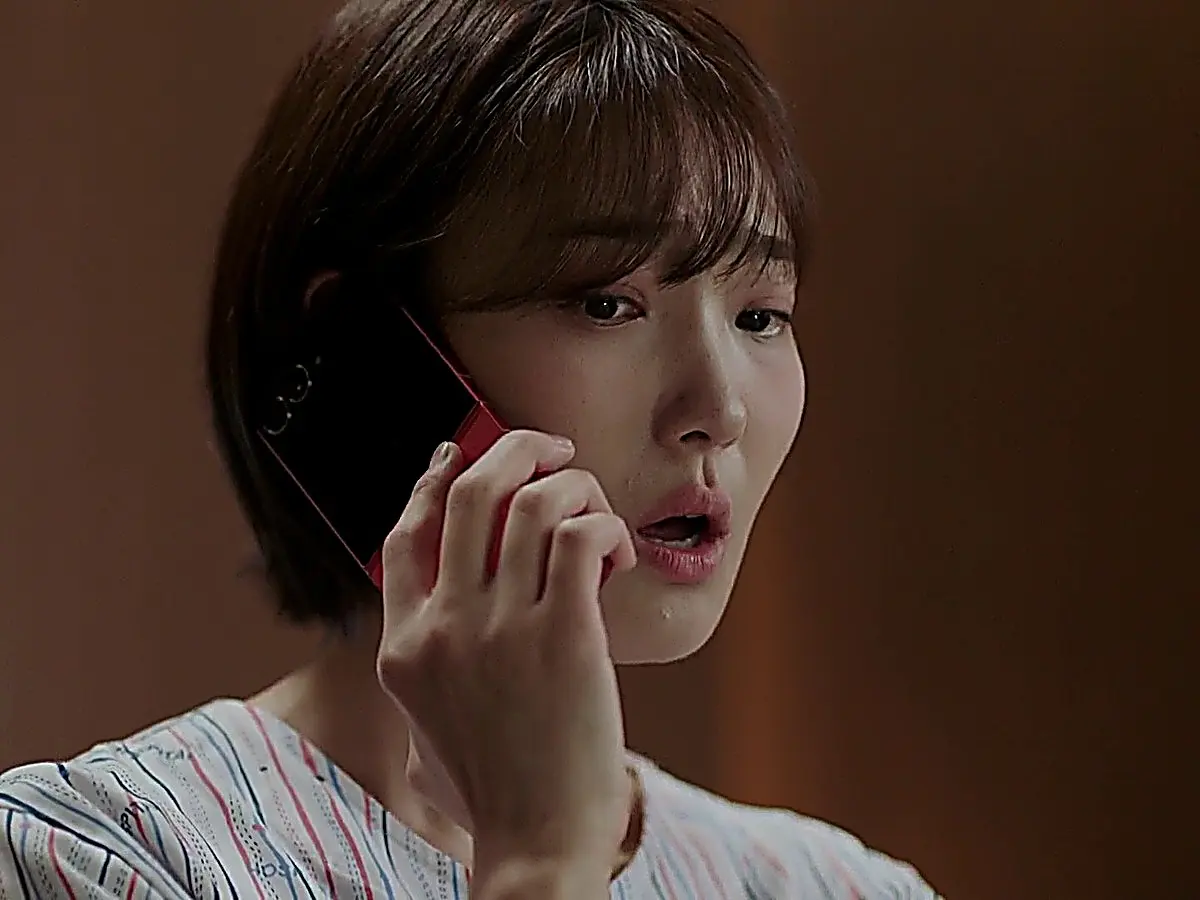 Seo-hyun holding a red flip phone to her ear while wearing a striped hospital gown