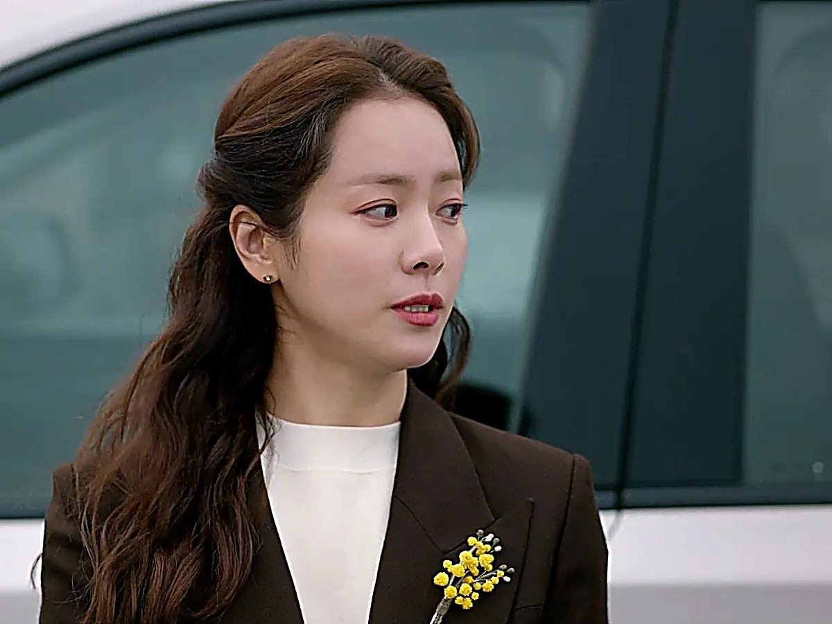 Lee Young-ok standing outside in a dark brown tailored blazer pinned with a small yellow floral brooch over a high-neck white top