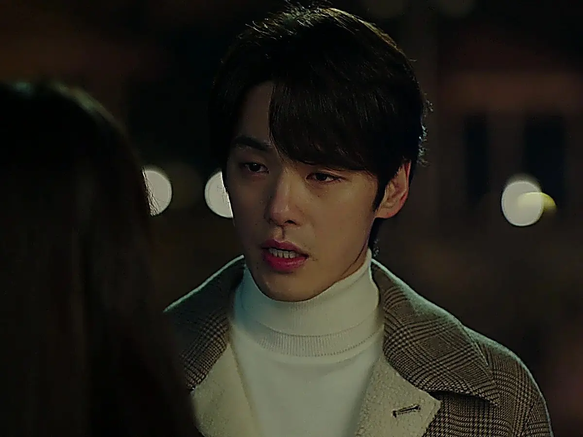 A man wearing a houndstooth coat and ivory turtleneck speaking outdoors at night
