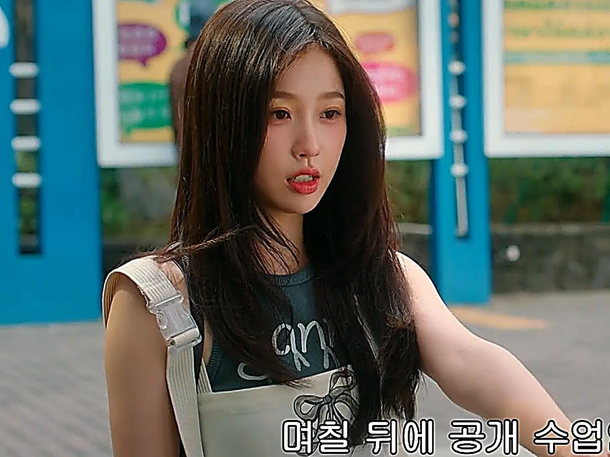 Jiang Mu with long dark hair wearing a dark green tank top under an off-white apron dress looking forward