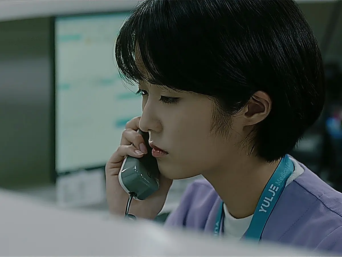 A medical staff member with short dark hair in purple scrubs holding a telephone receiver.