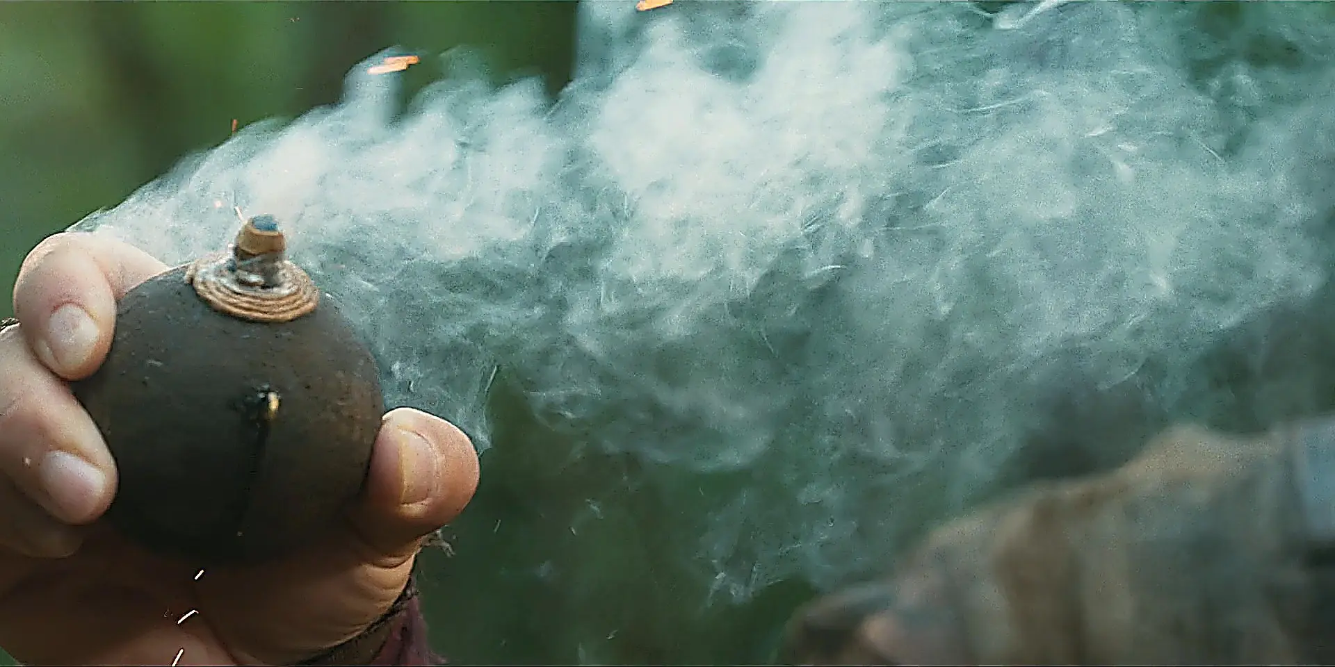 A close-up of a hand holding a spherical bomb emitting a thick plume of white smoke and flying sparks