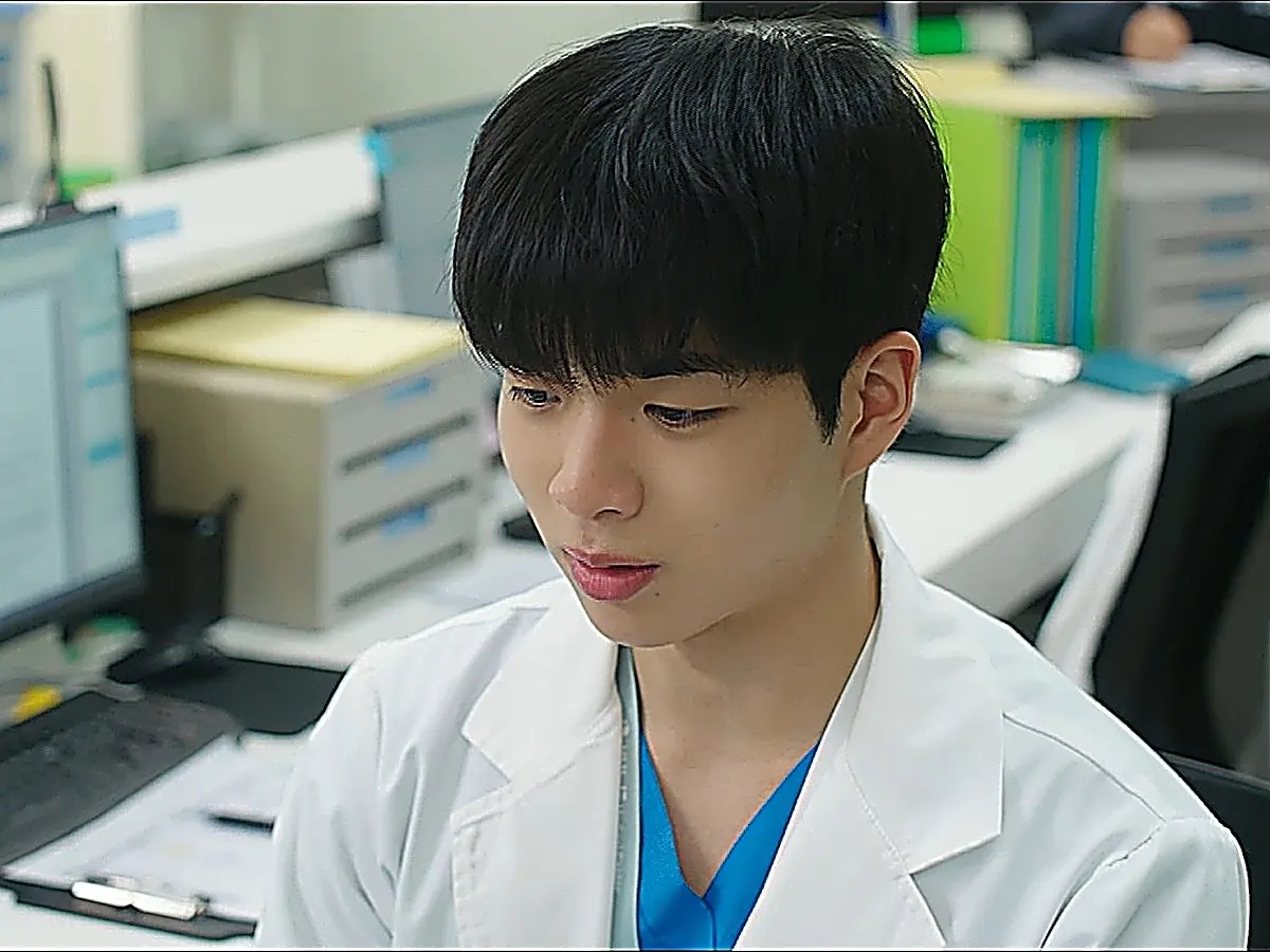A young man wearing a medical coat and scrubs looking down thoughtfully