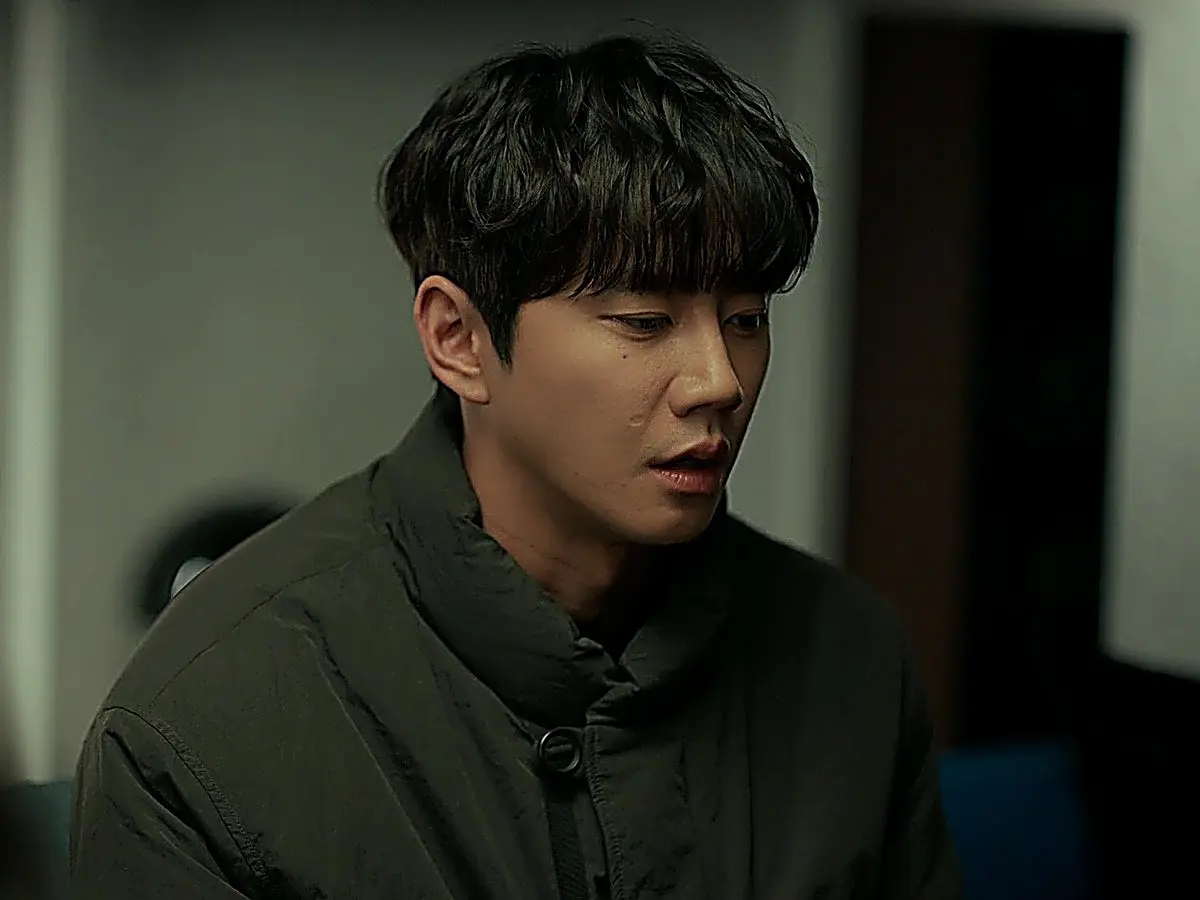 Man with textured dark hair in a dark padded coat looking down with a serious expression