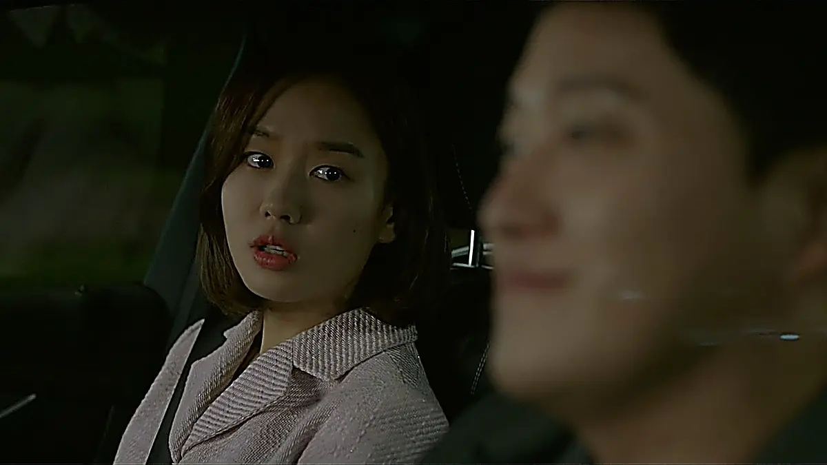 A woman with bobbed hair wearing a pink tweed jacket seated inside a vehicle at night.