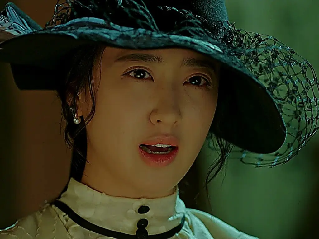 A woman wearing a black wide-brimmed hat with dark netting and a high-collared cream blouse looking forward