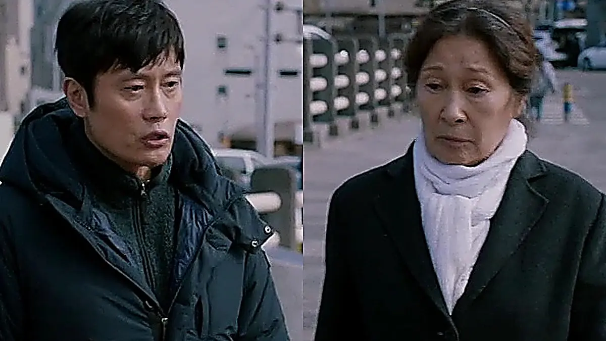 Lee Dong-seok in a black hooded down jacket over a dark zip-up fleece walking next to Kang Ok-dong dressed in a dark wool overcoat and white scarf