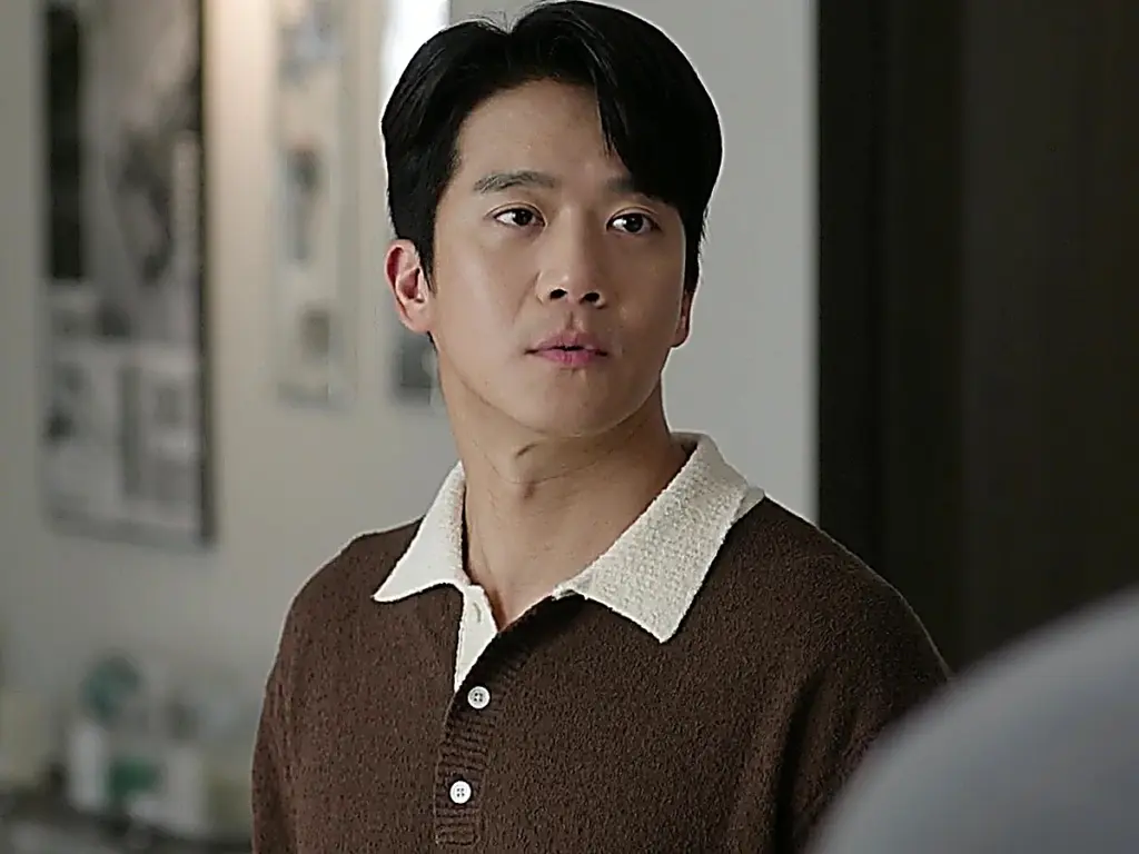 Mu-jin looking to the side in a brown knit polo shirt with a contrasting cream collar and white buttons