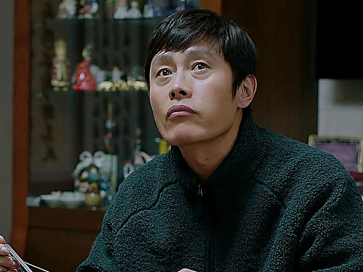Lee Dong-seok sitting at a dining table in a textured dark green sherpa fleece jacket with a high-neck zip collar