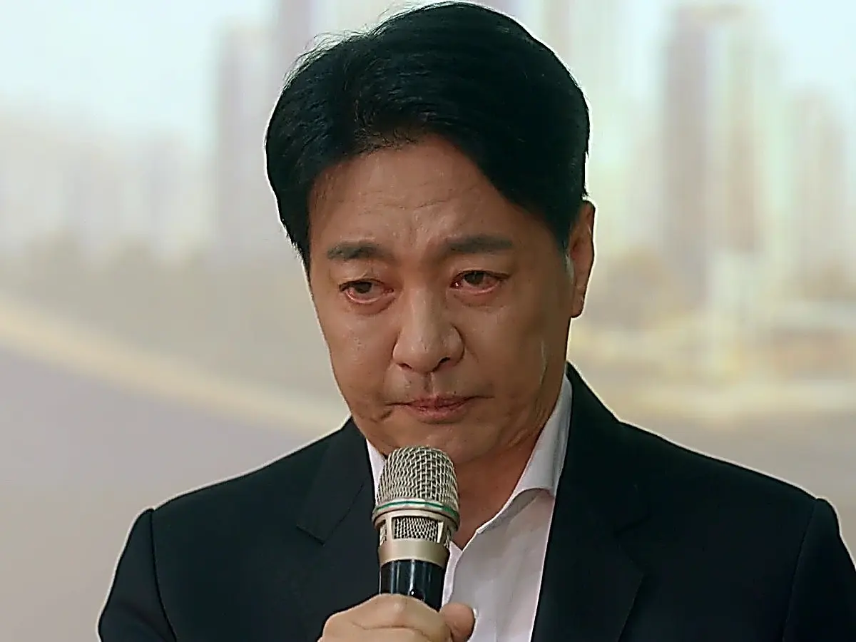 A man in a dark suit looking downward with a solemn face as he speaks into a microphone