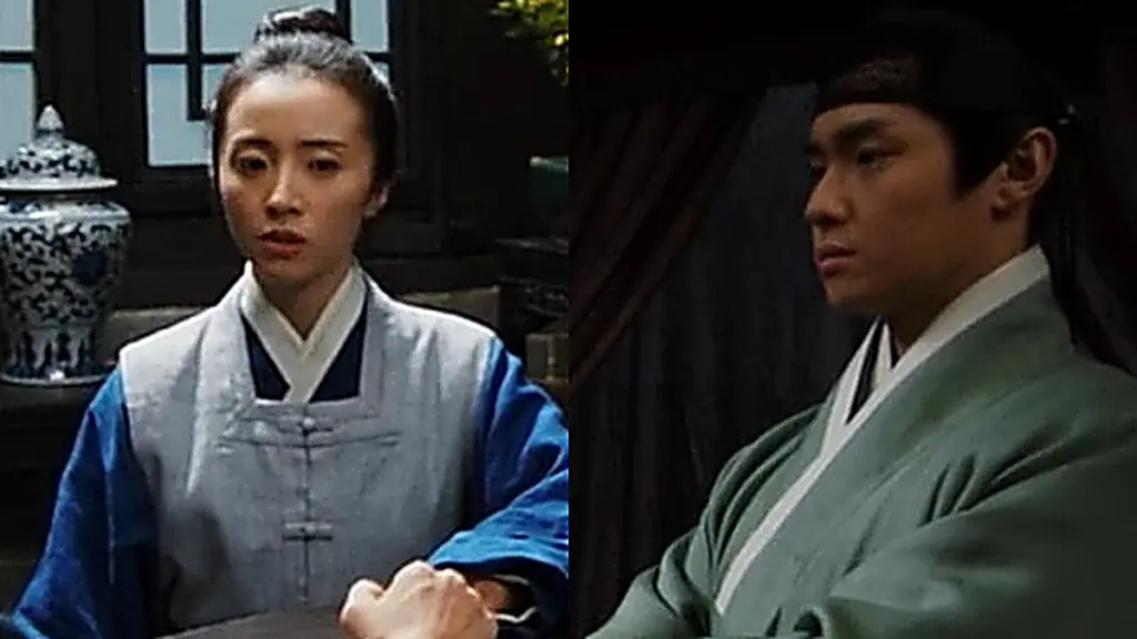 A woman in a grey vest and blue robe seated across a table from a man in a green robe