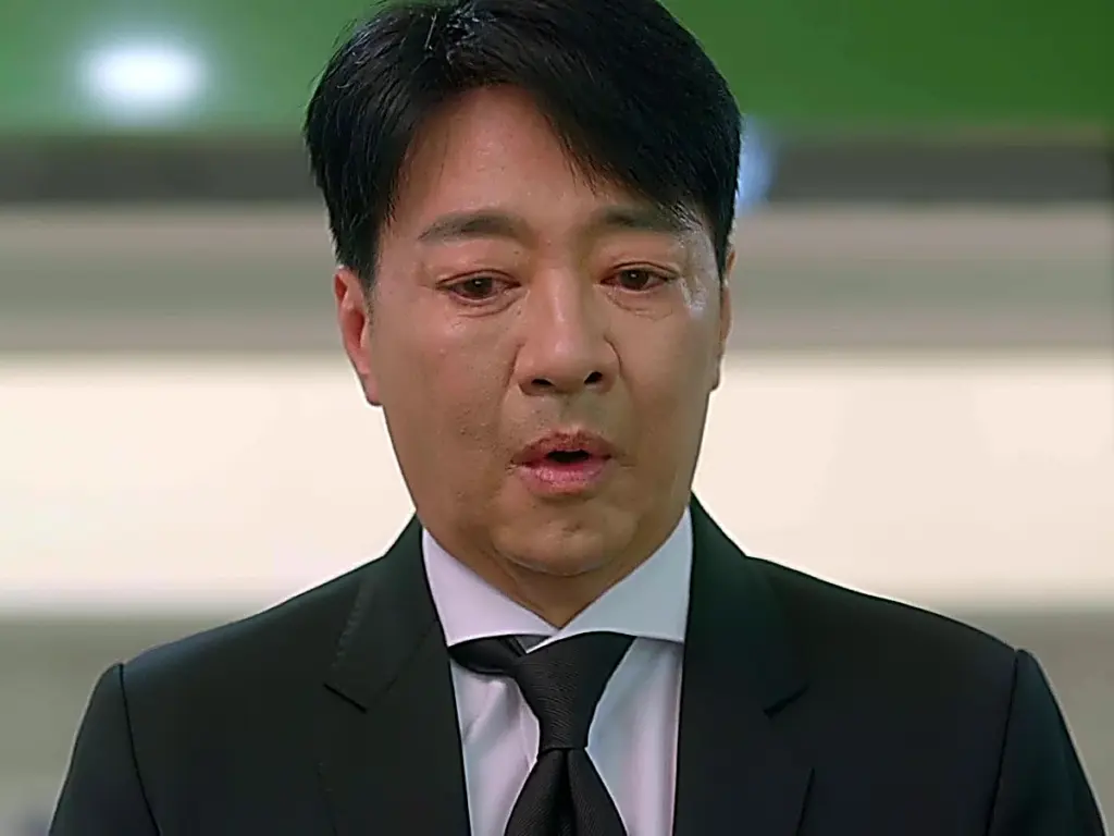 A man in a dark suit and black necktie looking downward with teary eyes
