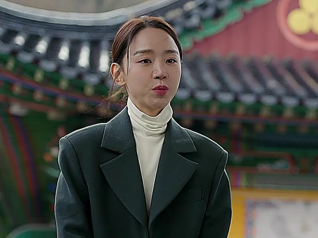 A woman in a dark green tailored coat and cream turtleneck standing outdoors in front of temple architecture