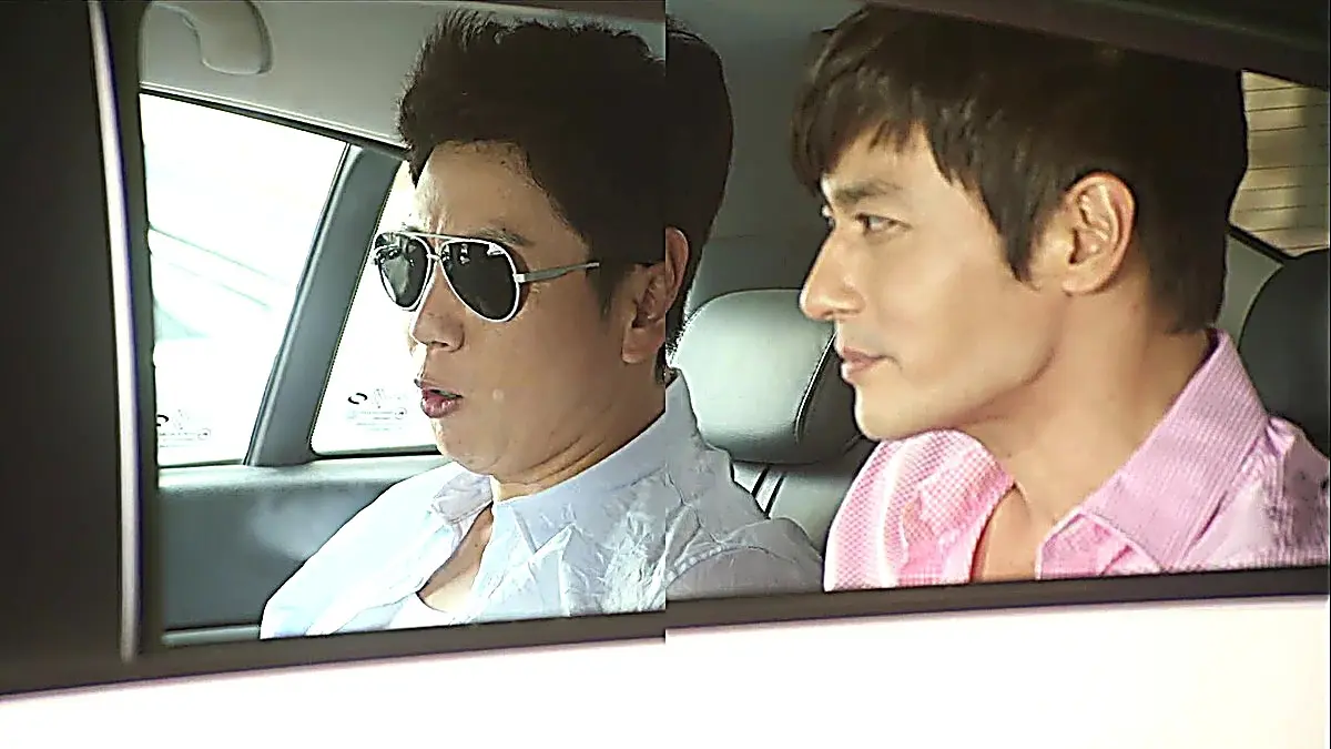 Two men seated in a car, one wearing sunglasses and a light blue collared shirt and the other in a pink checked button-up shirt