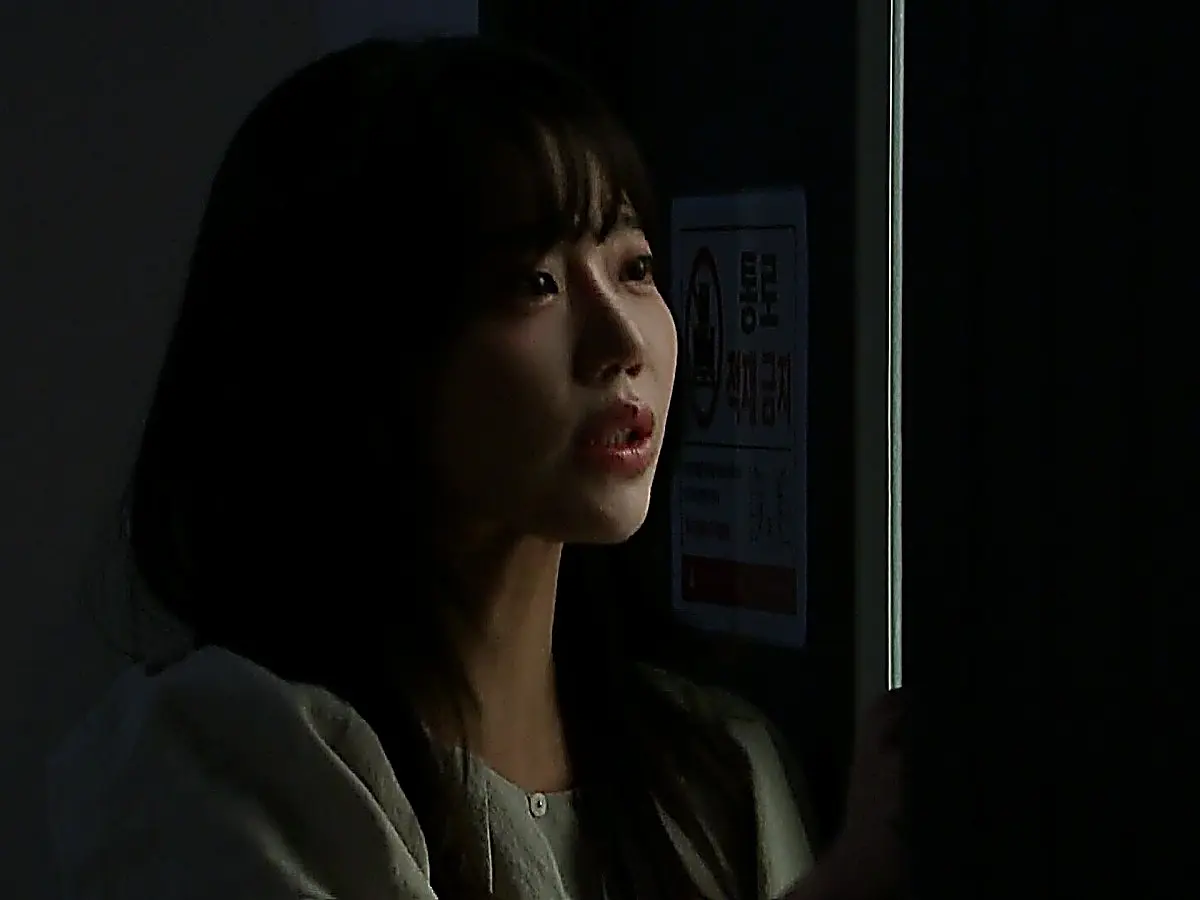 A woman in a dimly lit room peering out through a narrow gap in a dark doorway.