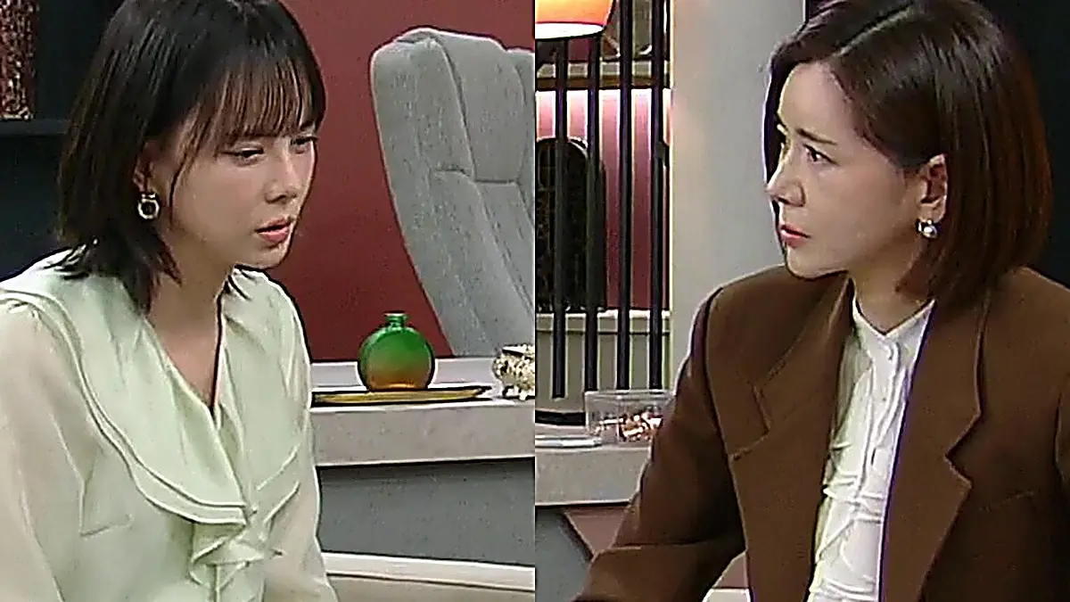 Two women facing each other across an office desk, one in a pale green blouse and the other in a brown blazer.
