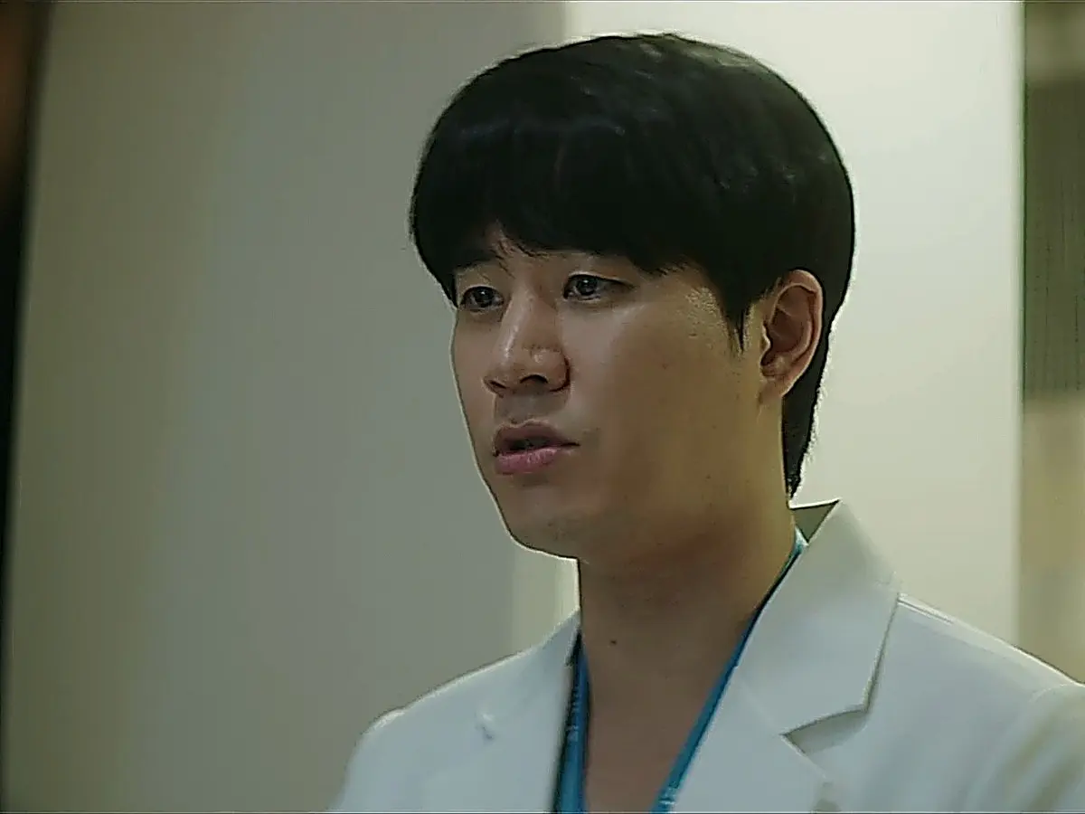 A doctor in a white lab coat over blue scrub top looking forward.