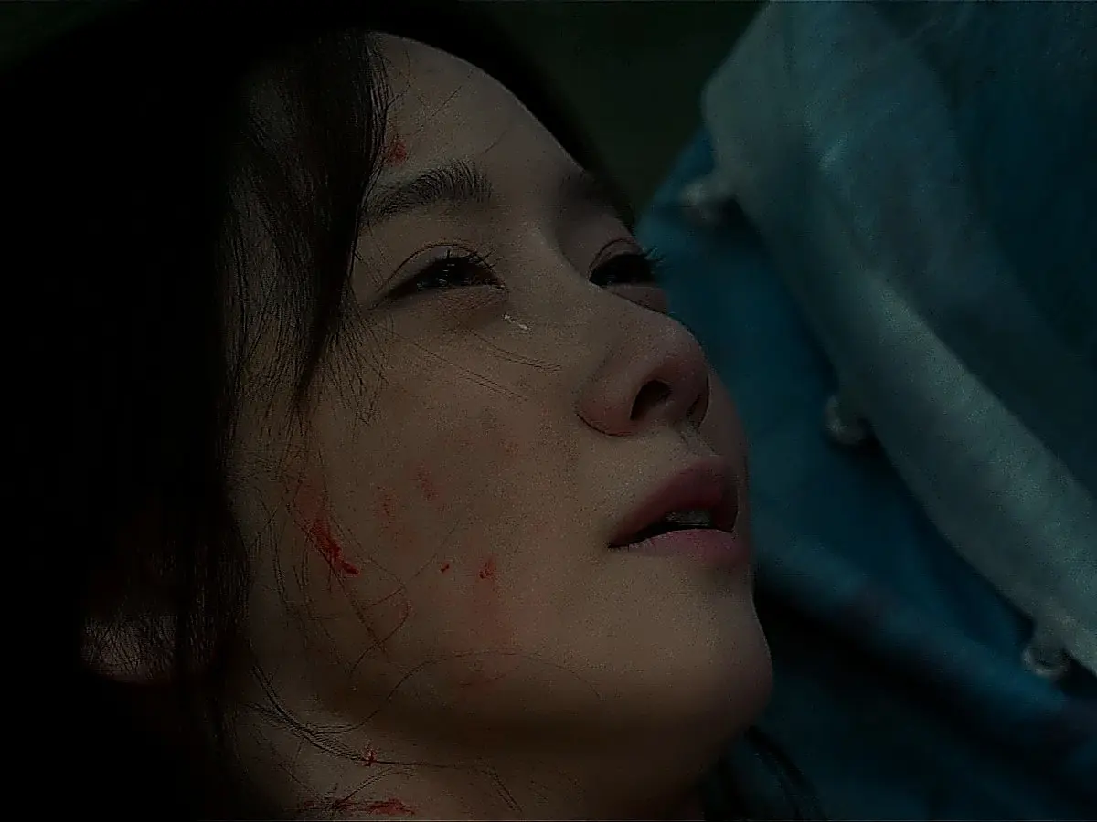 A wounded woman with red blood smears on her face looking upward while resting against blue fabric