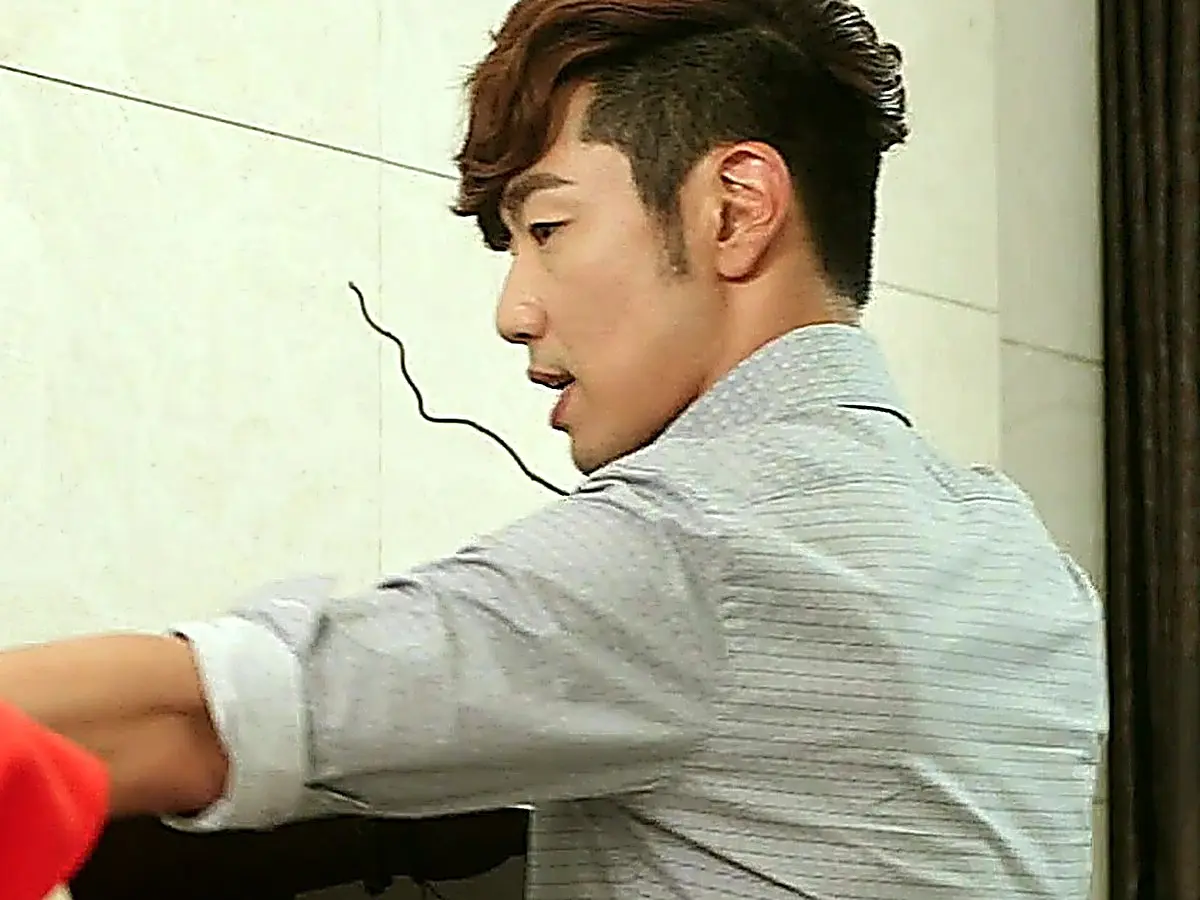 A man in a grey collared shirt seen from the side raising his arm in front of a pale wall