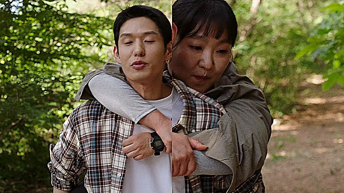 A man wearing a plaid button-up shirt carrying a woman in an olive shirt on his back along a wooded outdoor path.