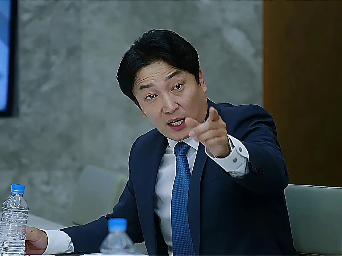 A man in a navy suit and blue patterned tie seated at a conference table pointing his finger forward
