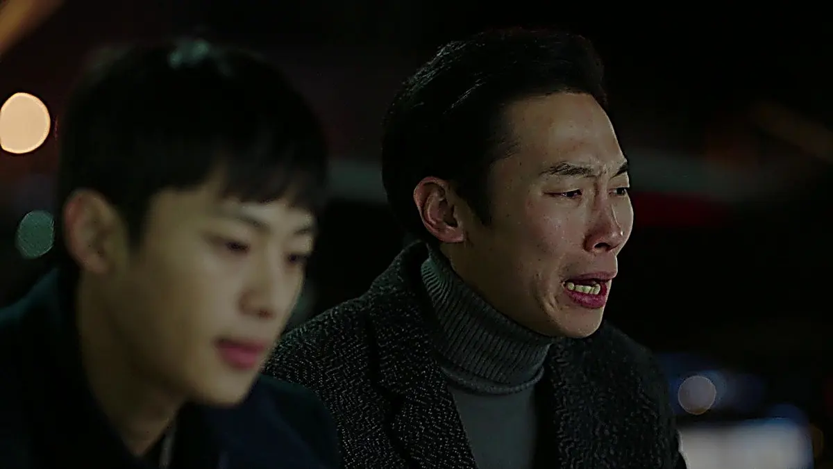 Two men sitting inside a vehicle at night, with one man grimacing in distress
