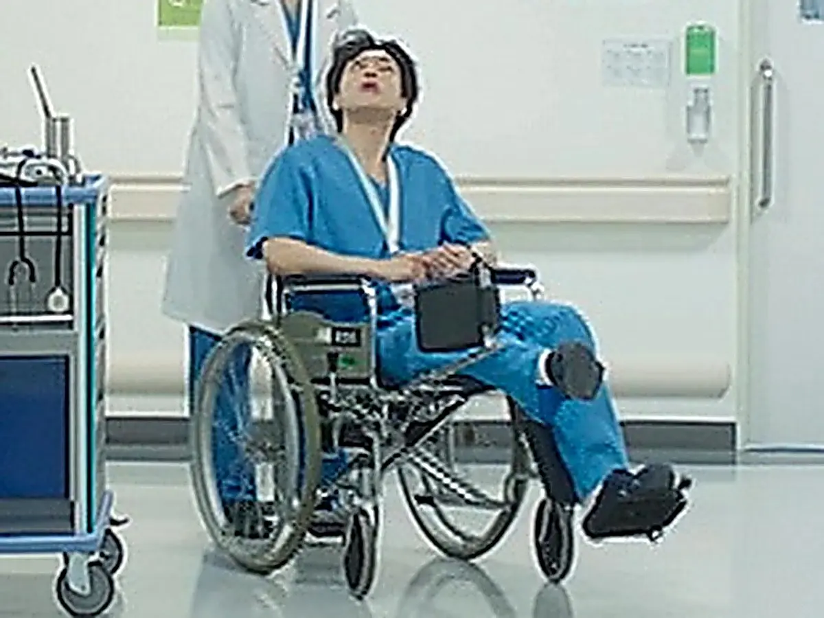 A person in blue scrubs seated in a wheelchair pushed along a hospital corridor