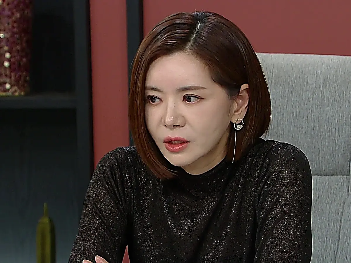 A woman with short brown bobbed hair wearing a black textured top looks forward with a stern expression