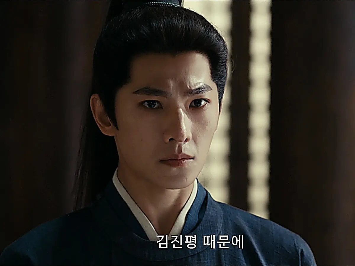 A man with tied-back dark hair wearing a deep blue collared robe looking forward