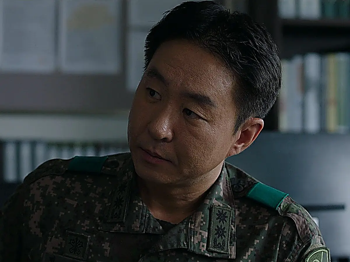 A man in a military digital camouflage uniform seated at an office desk looking toward the side.