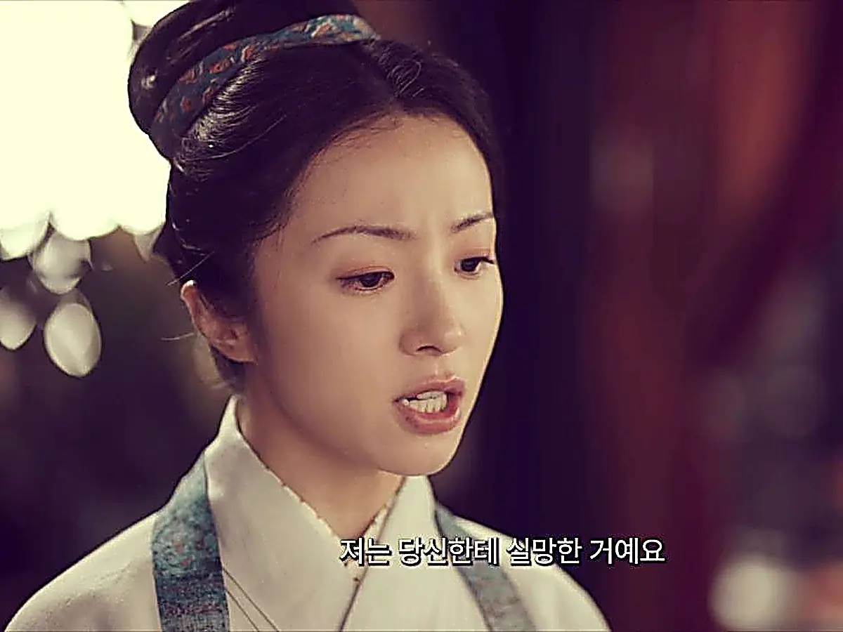 A woman with an updo hairstyle in an ivory robe speaking with a stern expression