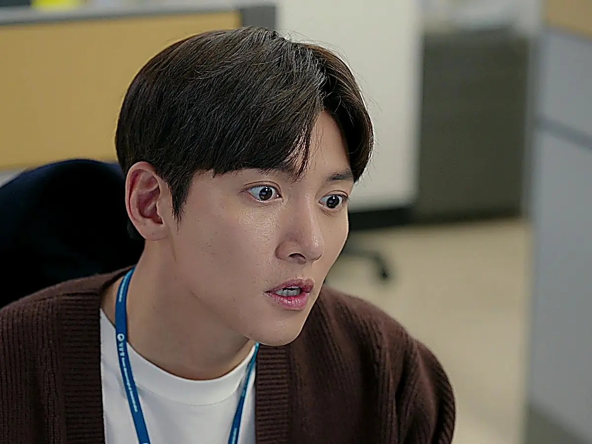 A young man wearing an office lanyard and a brown cardigan opening his eyes wide in sudden alarm