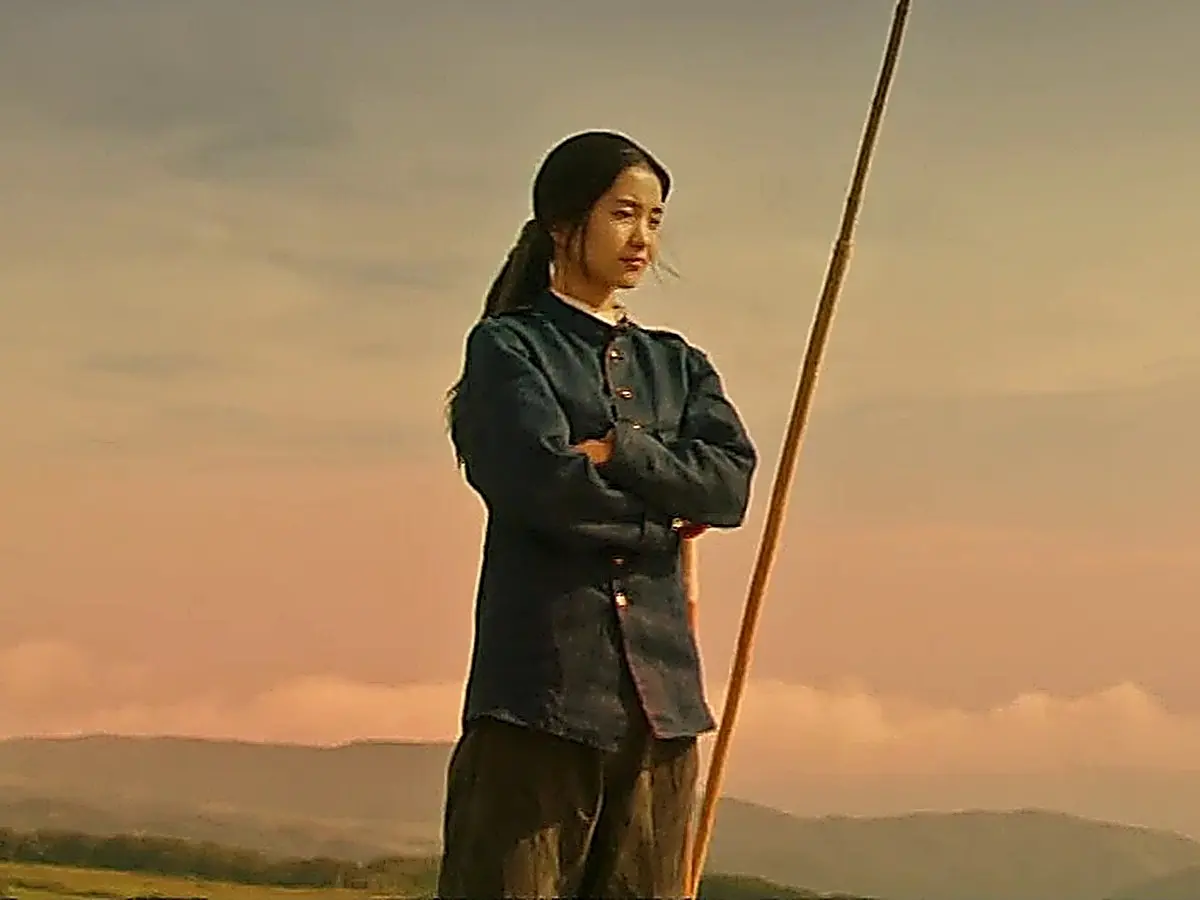 A woman with tied-back hair standing with crossed arms on an open field at sunset next to a tall pole